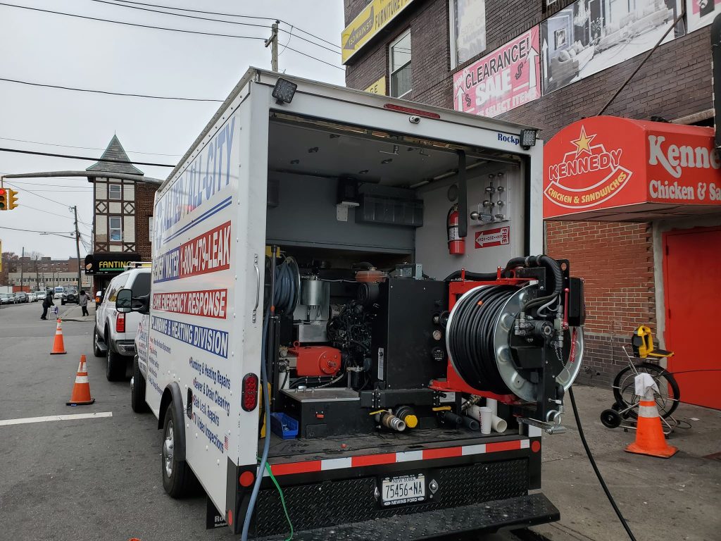 A service truck with its rear doors open reveals industrial cleaning equipment and hoses. It's parked on a city street near a Kennedy Chicken & Sandwiches restaurant and other storefronts.