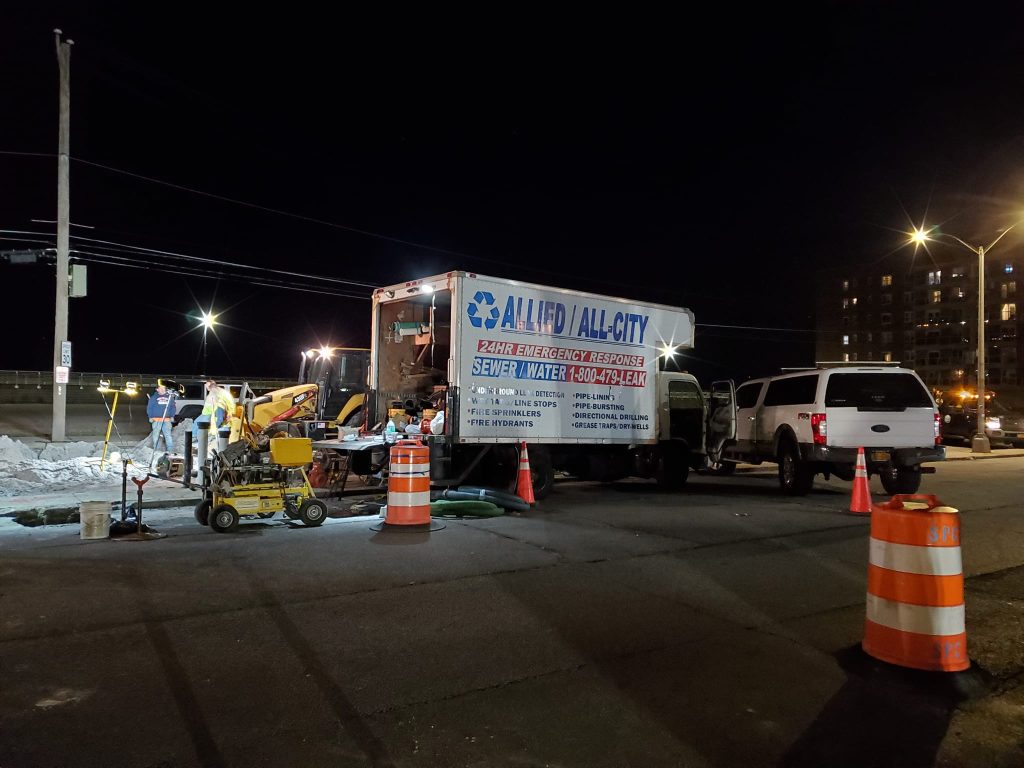 A nighttime street repair scene with workers, a backhoe, and equipment next to an "Allied All-City" emergency response truck. Orange traffic cones and barrels block off the area as sewer services are underway.