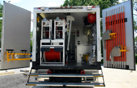 The back of a utility truck with its doors open, revealing equipment such as hoses, reels, tools, and machinery organized inside for maintenance or repair work.