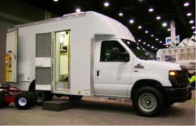 A white utility van with an open side door is parked indoors. Steps lead up to the entrance, revealing part of the van’s interior. The surrounding area appears to be an exhibition or trade show.