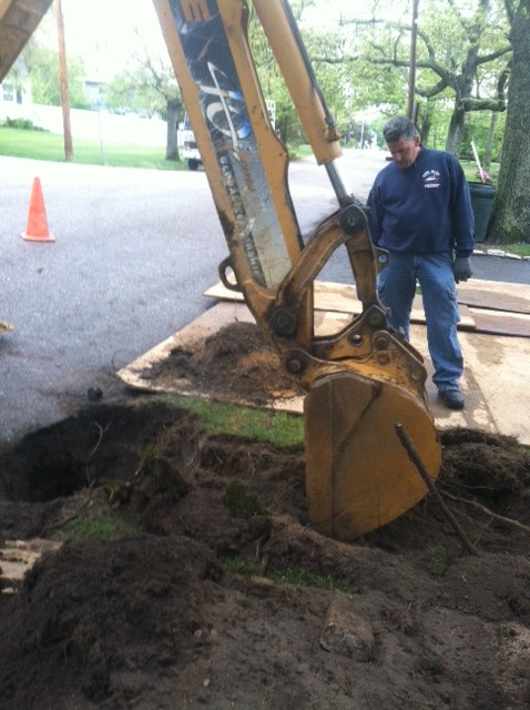 A man stands next to a yellow excavator digging a hole in the ground on a paved surface near grass, with an orange traffic cone in the background.