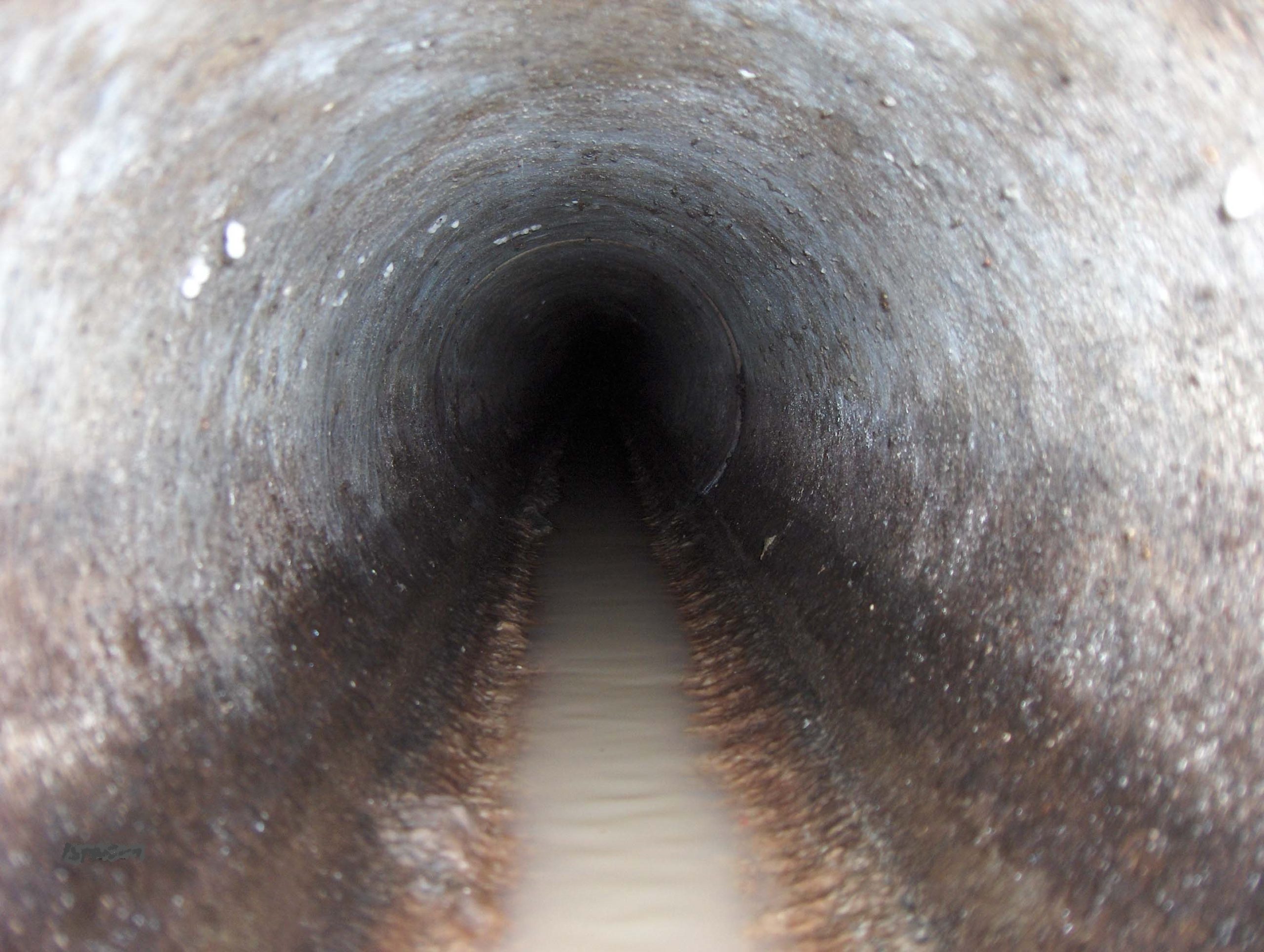 A close-up view inside a dark, cylindrical tunnel with wet, stained walls and a narrow stream of murky water running along the bottom.