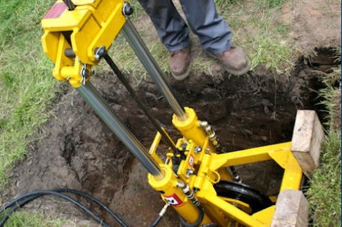 A person stands next to a yellow hydraulic machine positioned over a hole in the ground, with wooden blocks supporting the sides. The machine appears to be used for underground pipe installation or soil excavation.