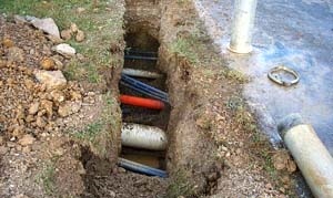 A trench dug in the ground with multiple utility pipes and cables, including black, orange, and white ones, exposed beneath the soil next to a paved road.