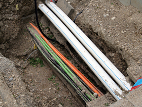 A close-up of several utility pipes and colorful cables, including orange, green, and black, laid side by side in a trench dug into the ground, surrounded by dirt and concrete.