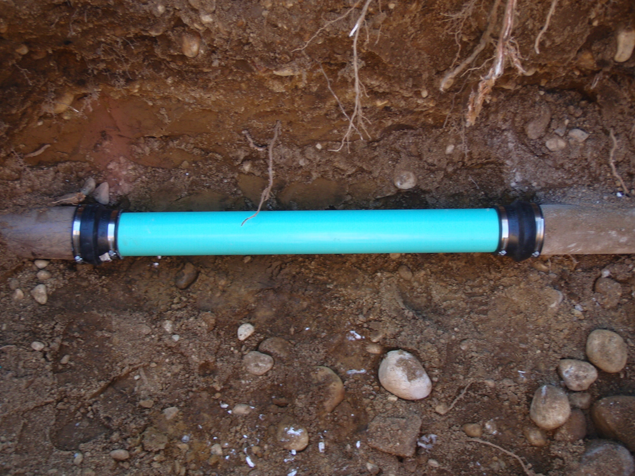 A section of blue PVC pipe has been used to repair a buried pipe, with metal couplings on both ends, surrounded by soil and rocks in a trench.