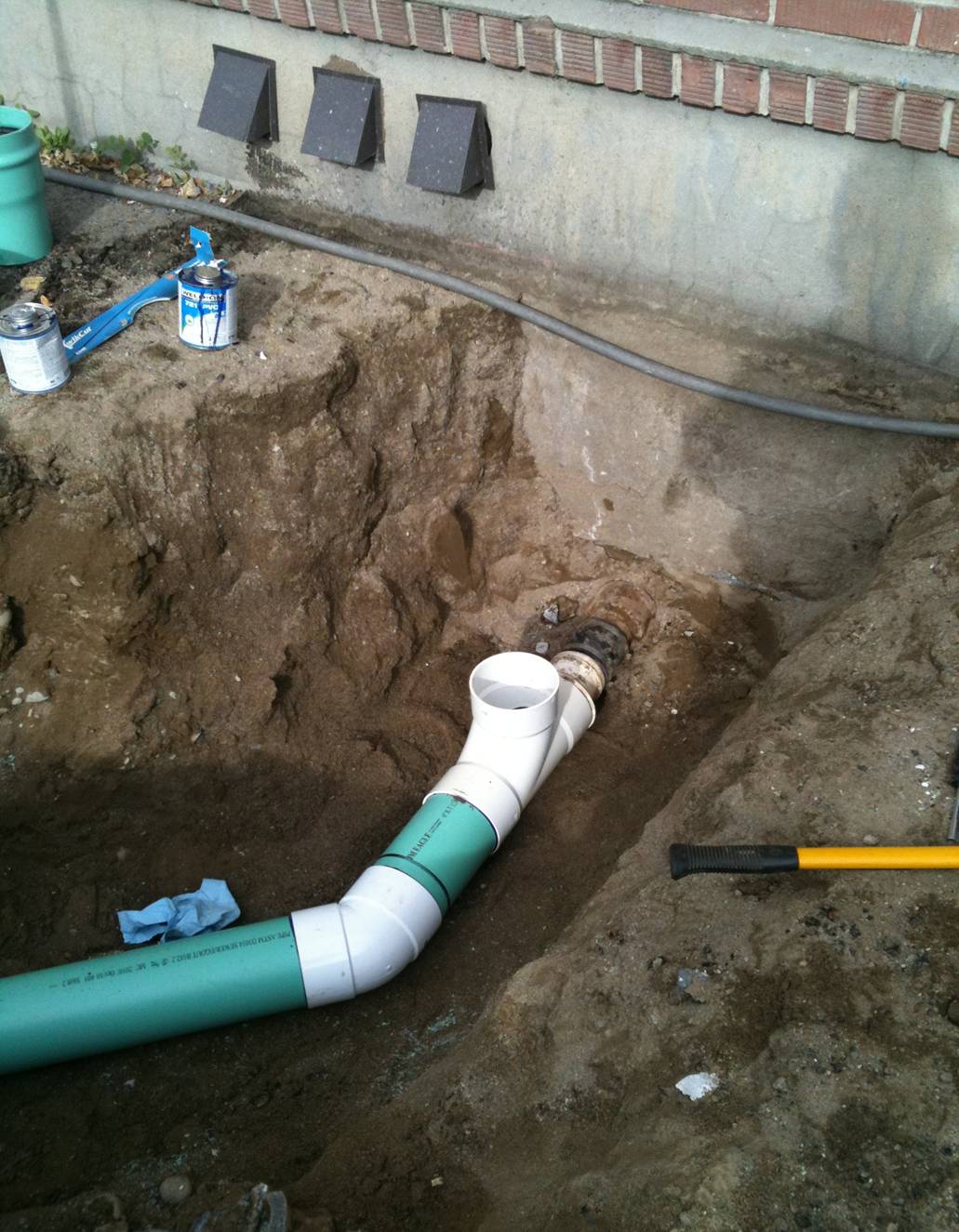 A section of green and white PVC plumbing pipe installed in a dirt trench next to a concrete foundation wall, with tools and supplies nearby.
