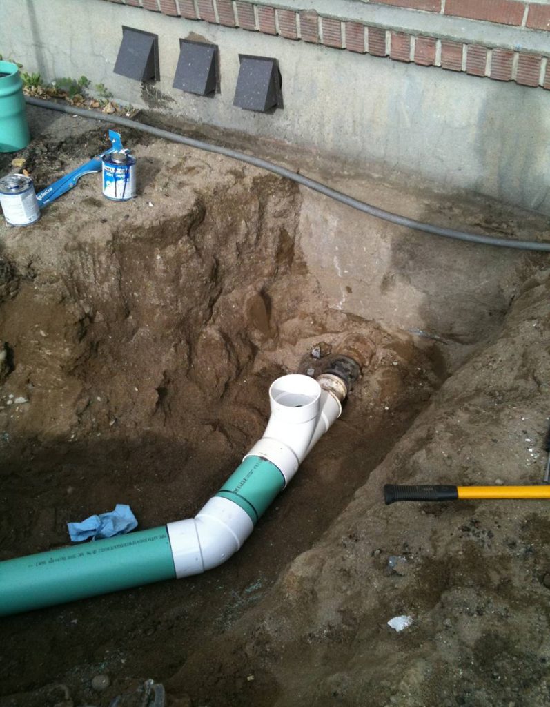 A partially buried green and white PVC sewer pipe installation next to a building foundation, with exposed soil, tools, and plumbing supplies nearby.