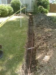 A narrow trench dug through a grassy yard with a pipe or cable running along the bottom, soil piled on one side, and yellow caution tape marking the area.