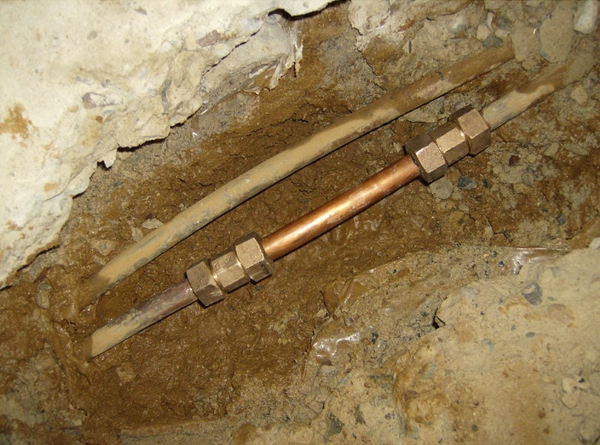 A close-up of a copper pipe repair with brass compression fittings connects two sections of pipe, partially buried in muddy soil within a dug-out area, showcasing expert water line services.