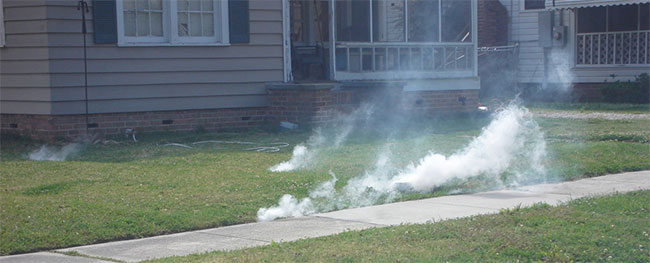 White smoke is rising from a grassy area beside a sidewalk in front of a house, suggesting possible fumigation or a small outdoor fire. Nearby, another house is visible in the background.