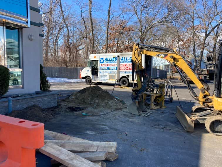 A small excavator is digging near a pile of dirt on a paved surface, with wooden planks and orange barriers nearby. A moving truck with "Allied" branding is parked in the background near trees.