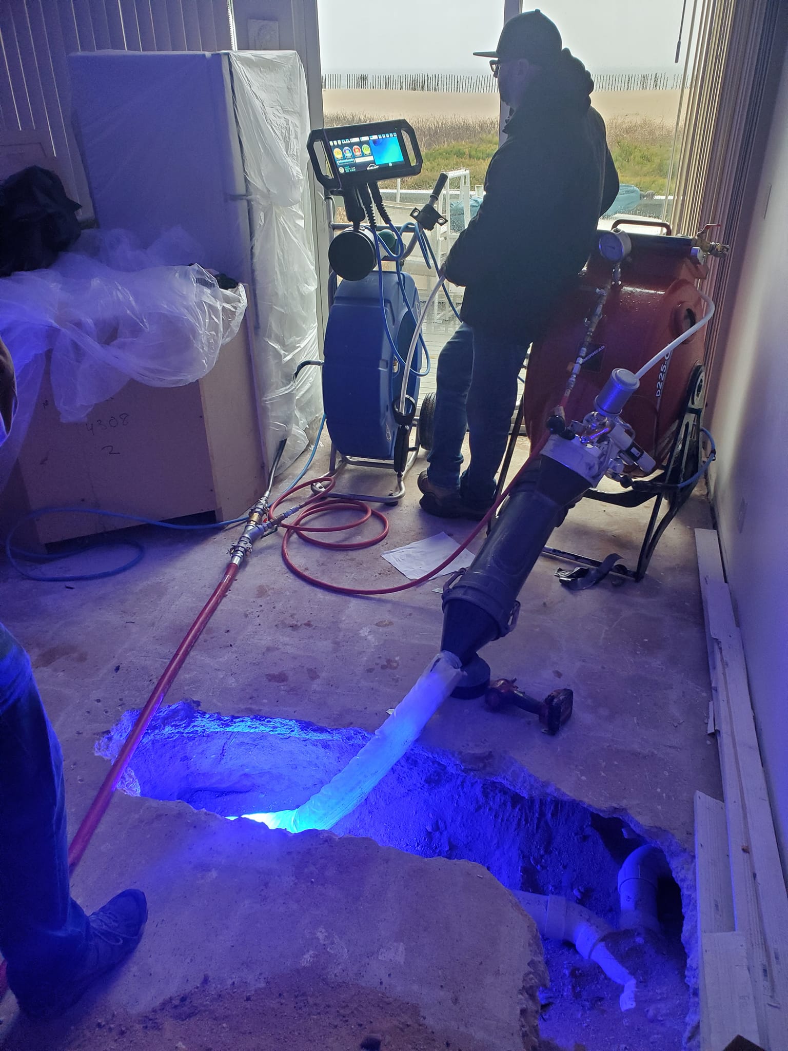 A worker operates specialized equipment by a large hole in a concrete floor, with blue light illuminating pipework inside the hole. Various tools, cables, and a machine fill the room.