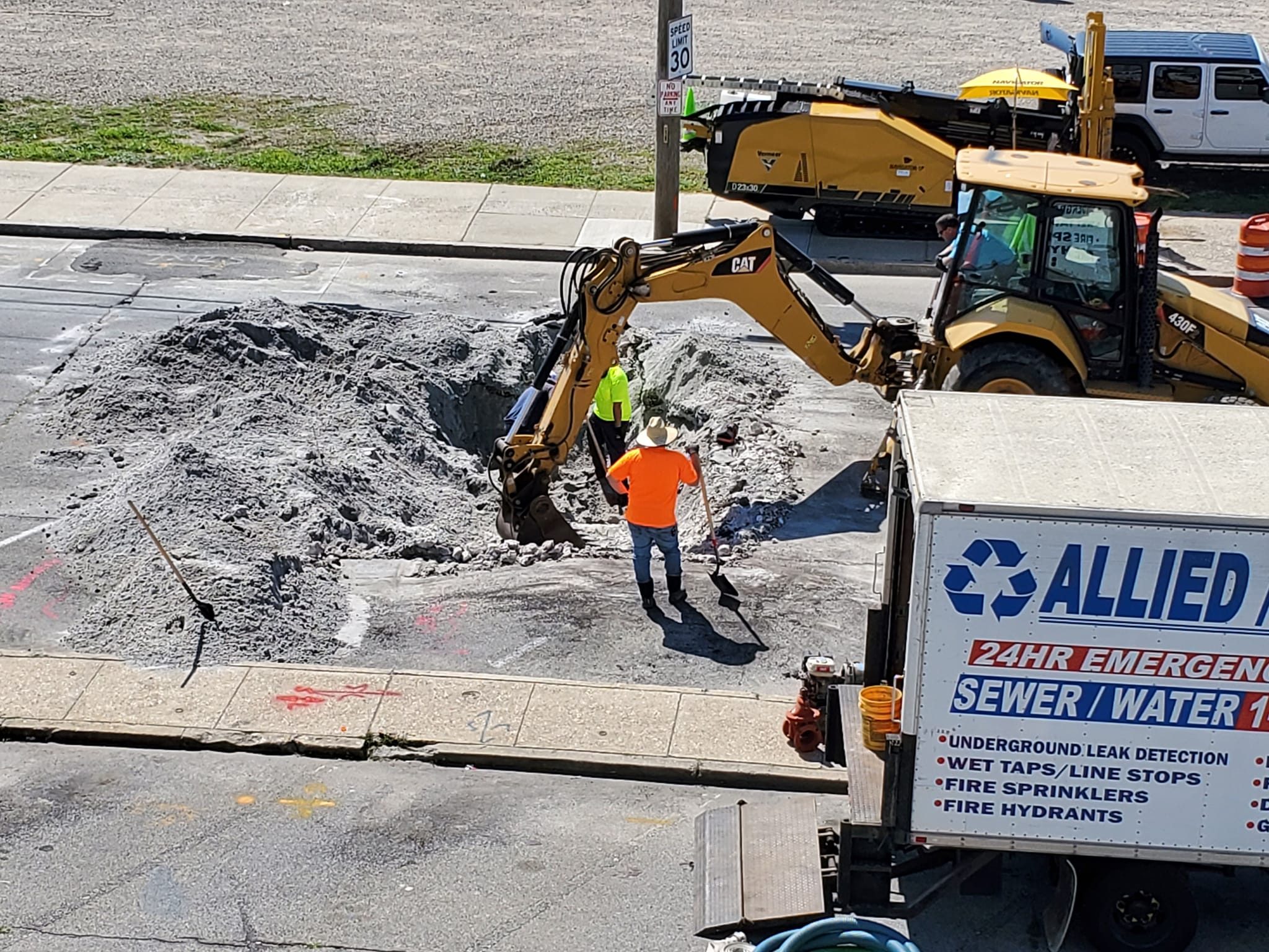 Two workers in safety vests use a backhoe to dig a large hole in a city street near a sewer repair truck. The area is covered in sand and construction equipment; traffic cones are nearby.