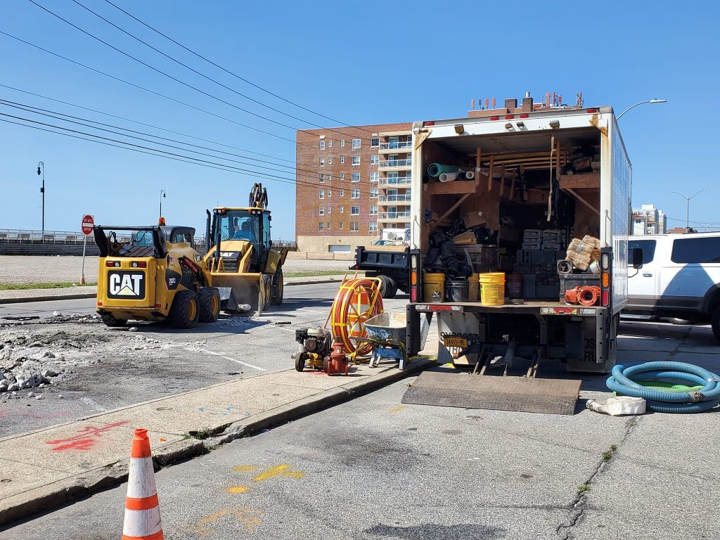 A construction site on a city street with a backhoe loader breaking pavement, an open utility truck with equipment, traffic cones, hoses, and a partially visible building in the background under a clear blue sky.