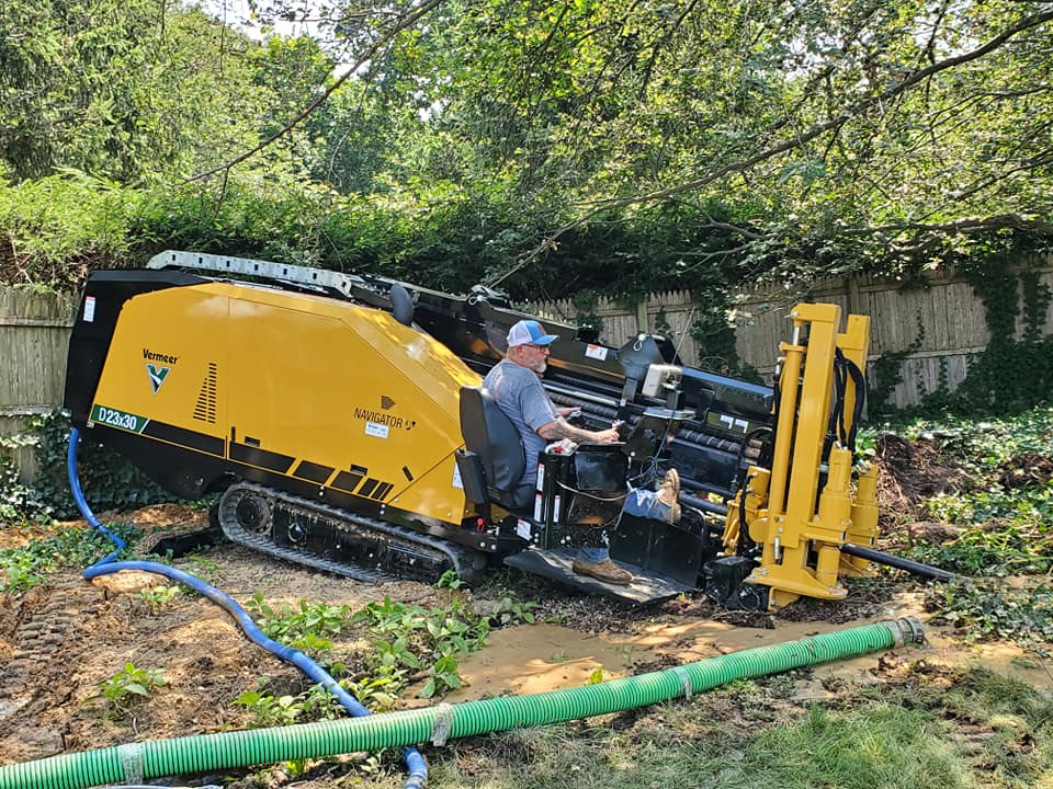 A man in a blue cap operates a large yellow Vermeer Navigator horizontal directional drill on muddy ground, surrounded by greenery and pipes, with a wooden fence in the background.