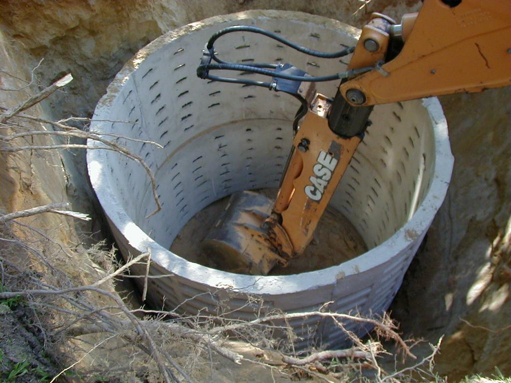 A CASE excavator places or adjusts a large concrete well ring in a round, dug-out pit, surrounded by soil and tree branches.