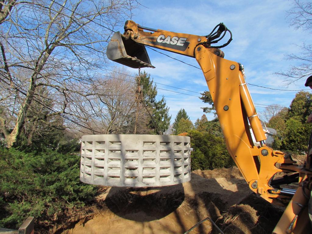 A yellow CASE excavator lifts a large concrete septic tank ring at a construction site surrounded by trees and bushes under a blue sky.