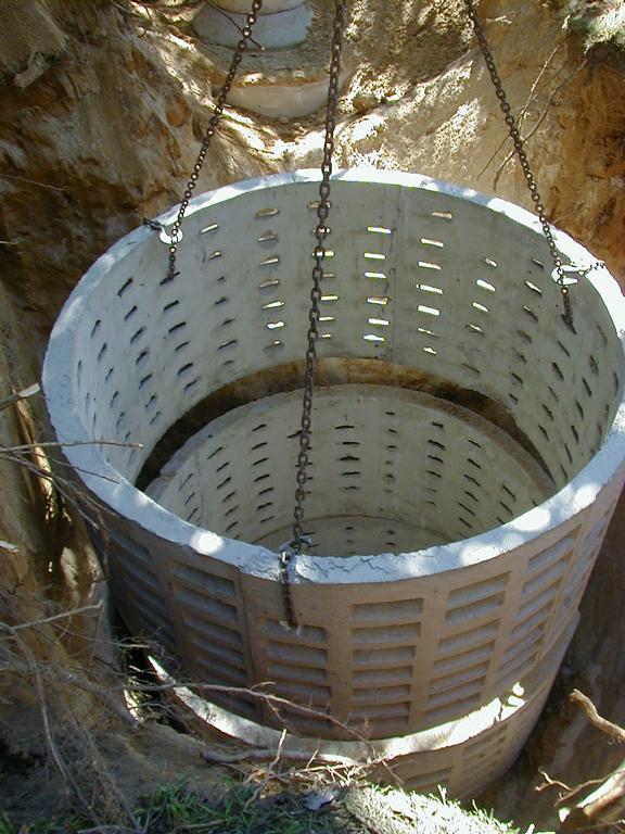 A large concrete drainage cylinder with rectangular holes is being lowered into a dug-out pit using metal chains. Sunlight illuminates the structure and the surrounding soil.