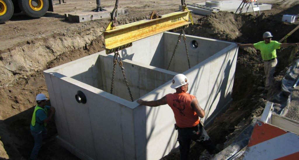 Three construction workers guide a large concrete utility box being lowered into a trench by a crane at a construction site. The workers wear safety vests, helmets, and work boots.