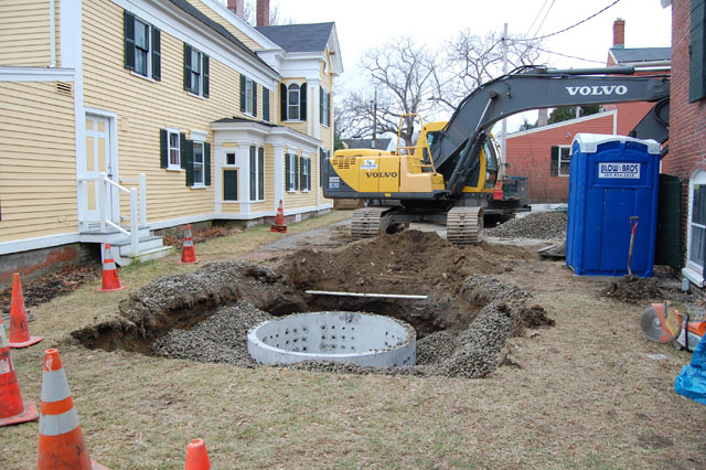 A construction site between two houses features an excavator, a large hole with a concrete cylinder inside, gravel, orange cones, and a portable toilet nearby.
