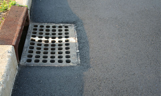 A close-up view of a storm drain with a metal grate and holes, set into freshly paved asphalt next to a concrete curb.