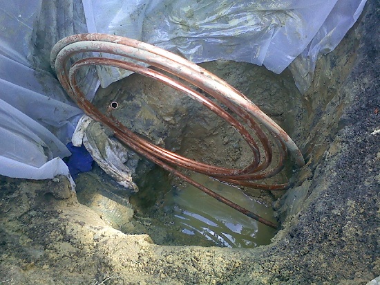A coiled copper pipe is partially submerged in muddy water in a shallow dug hole, with plastic sheeting lining the sides of the pit.