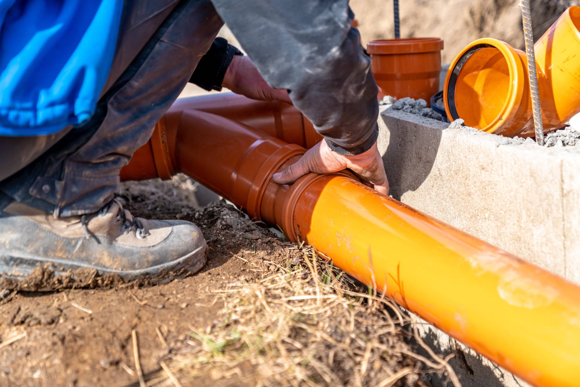 A worker in boots and a blue jacket is installing large orange plastic pipes outdoors, joining the pipe sections together near a concrete edge surrounded by soil and grass.