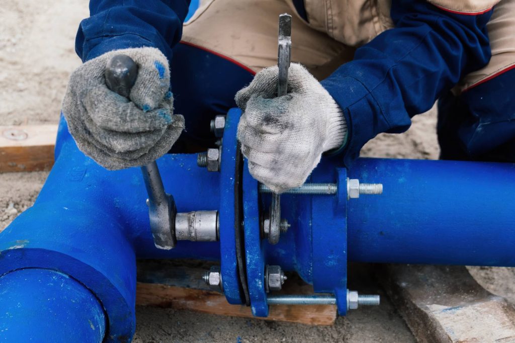 A person wearing gloves uses tools to tighten bolts on a large blue pipe joint, working outdoors on plumbing or pipeline installation.
