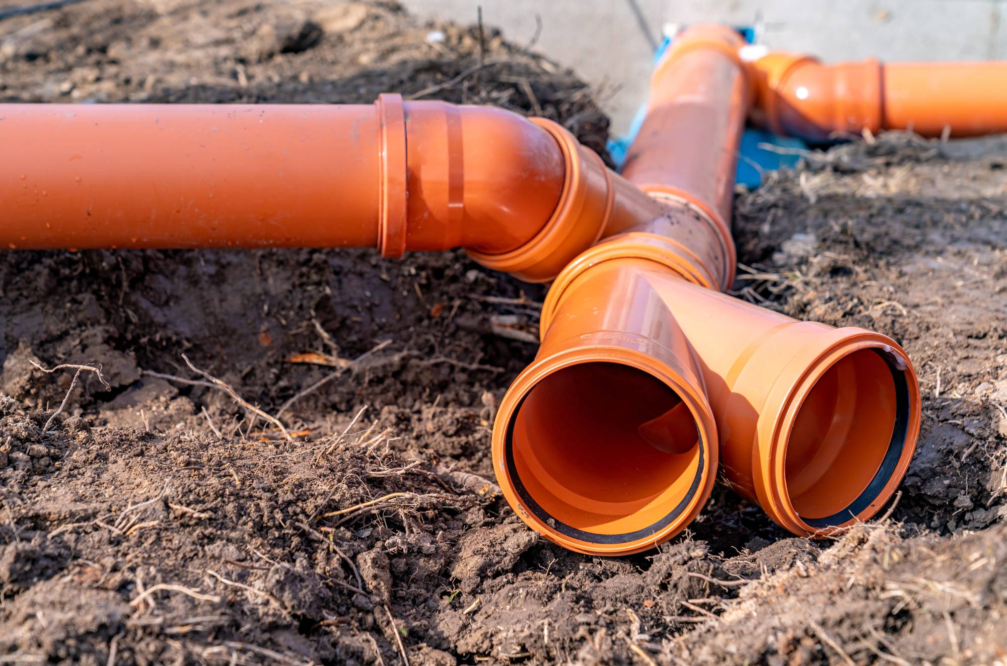 Close-up of orange PVC drainage pipes being installed in the ground, with several pipe joints and connectors visible, surrounded by soil and earth.