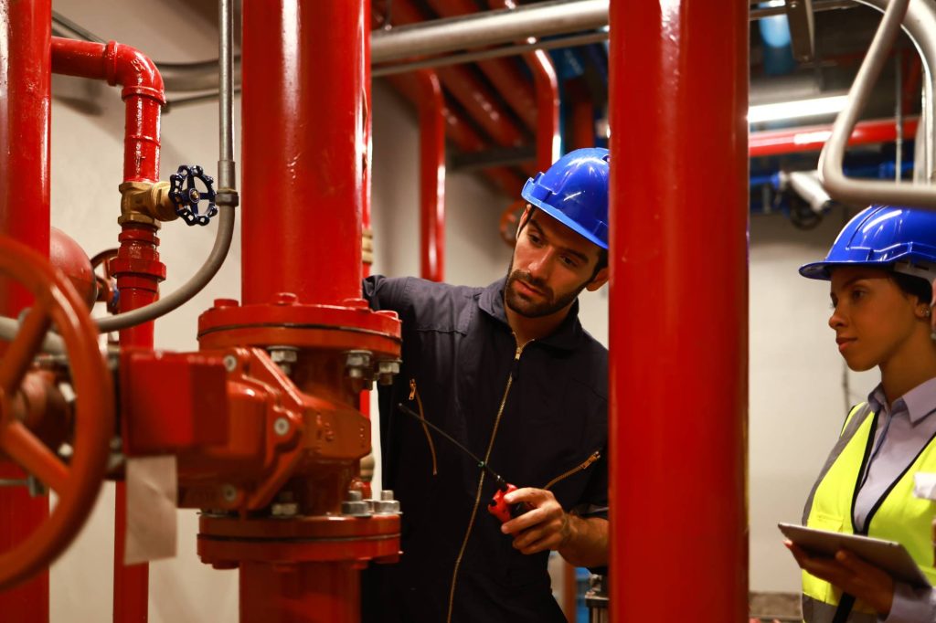Two engineers wearing blue safety helmets inspect and work on large red industrial pipes in a mechanical room; one holds tools and examines the pipes while the other takes notes on a clipboard.