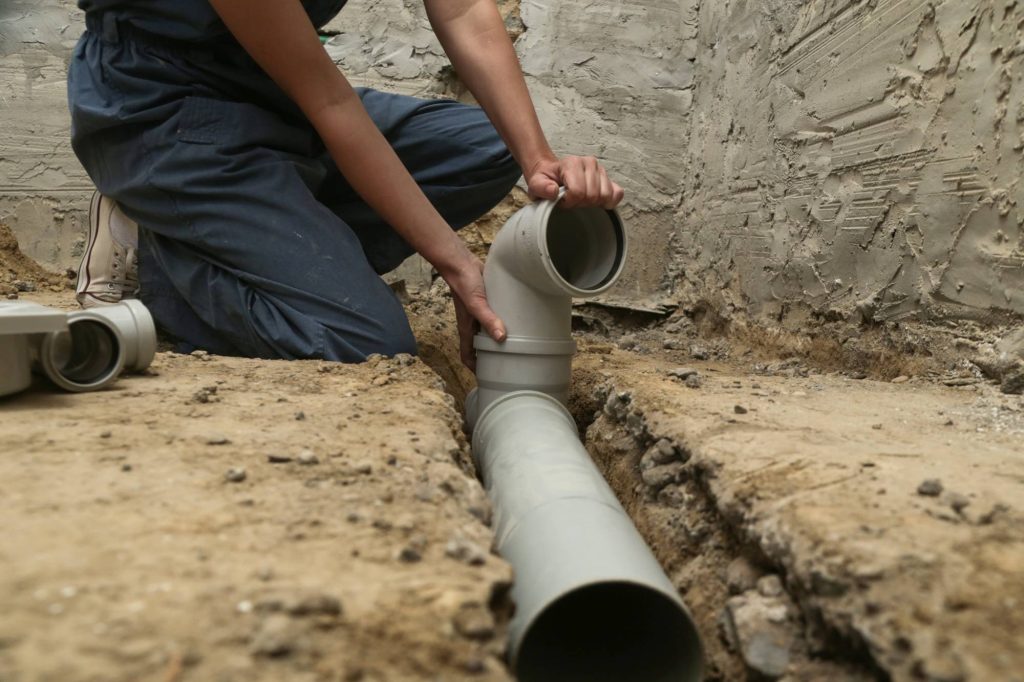 A person kneels on the ground, installing PVC plumbing pipes in a dirt trench within an unfinished, concrete-walled area.