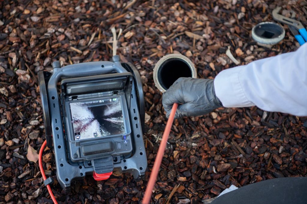 A person wearing a glove inserts a cable into an outdoor pipe while monitoring the view on a sewer inspection camera, surrounded by brown mulch and tools.