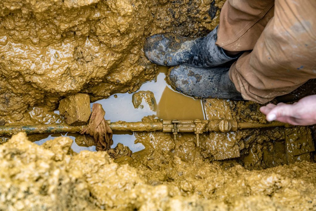 A person wearing muddy boots and brown pants stands in a muddy trench next to a pipe partially submerged in brown water. Their hand is visible near the pipe, which is surrounded by wet, packed earth.