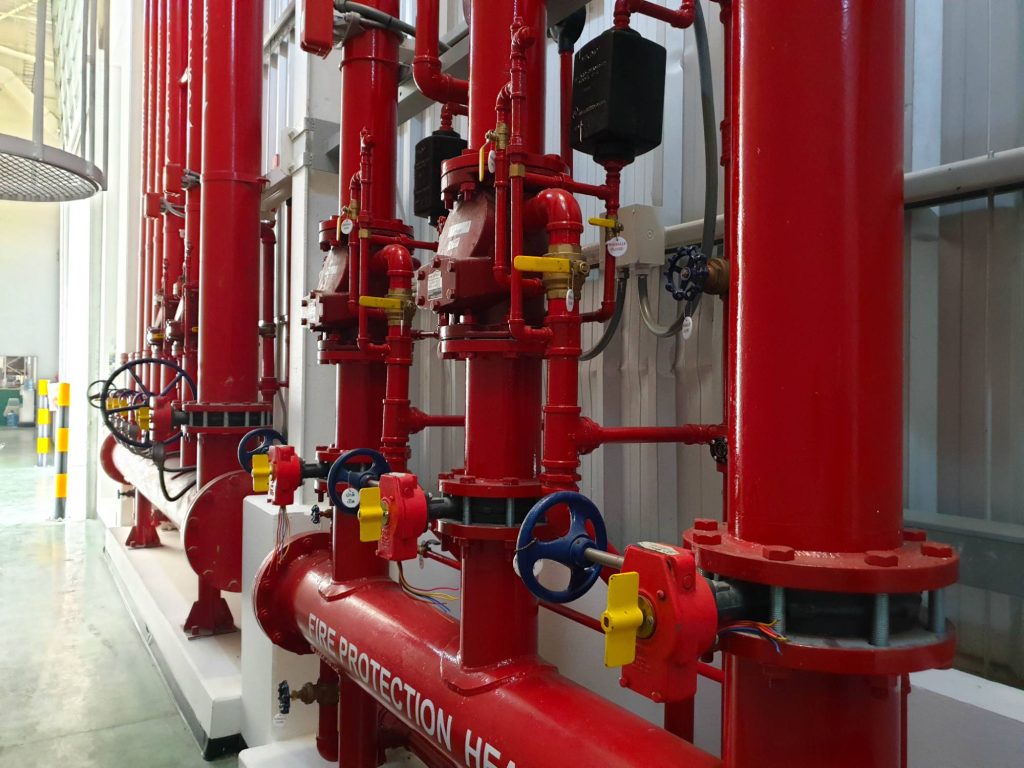 Rows of red fire protection pipes and valves in an industrial facility. Blue and yellow handwheels, gauges, and control boxes are visible, with the pipes running vertically and horizontally along a metallic wall.