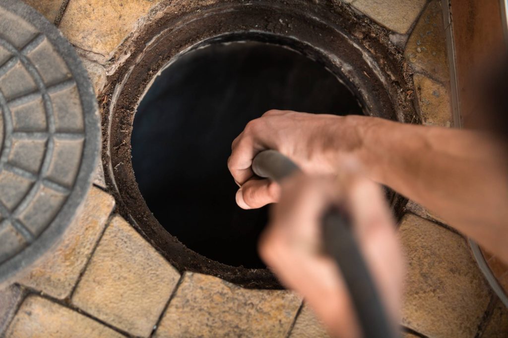 A hand holding a hose or pipe is seen above an open manhole surrounded by brown and tan tiles, suggesting maintenance or cleaning work.