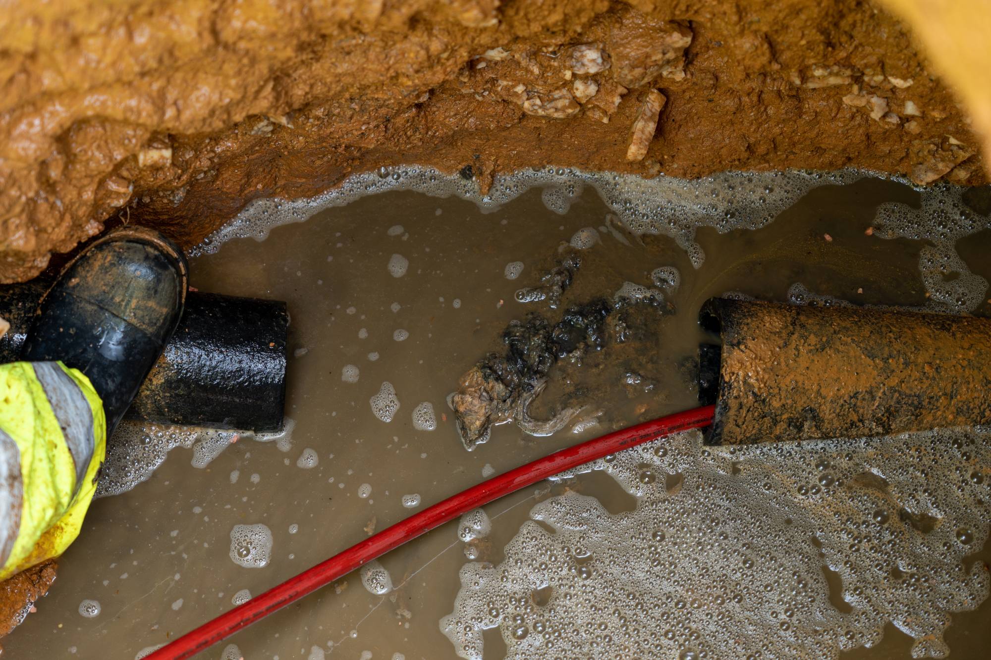 A muddy trench with water, two black pipes—one coated in mud—and a red cable submerged. Part of a worker's boot and high-visibility clothing is visible on the left side.