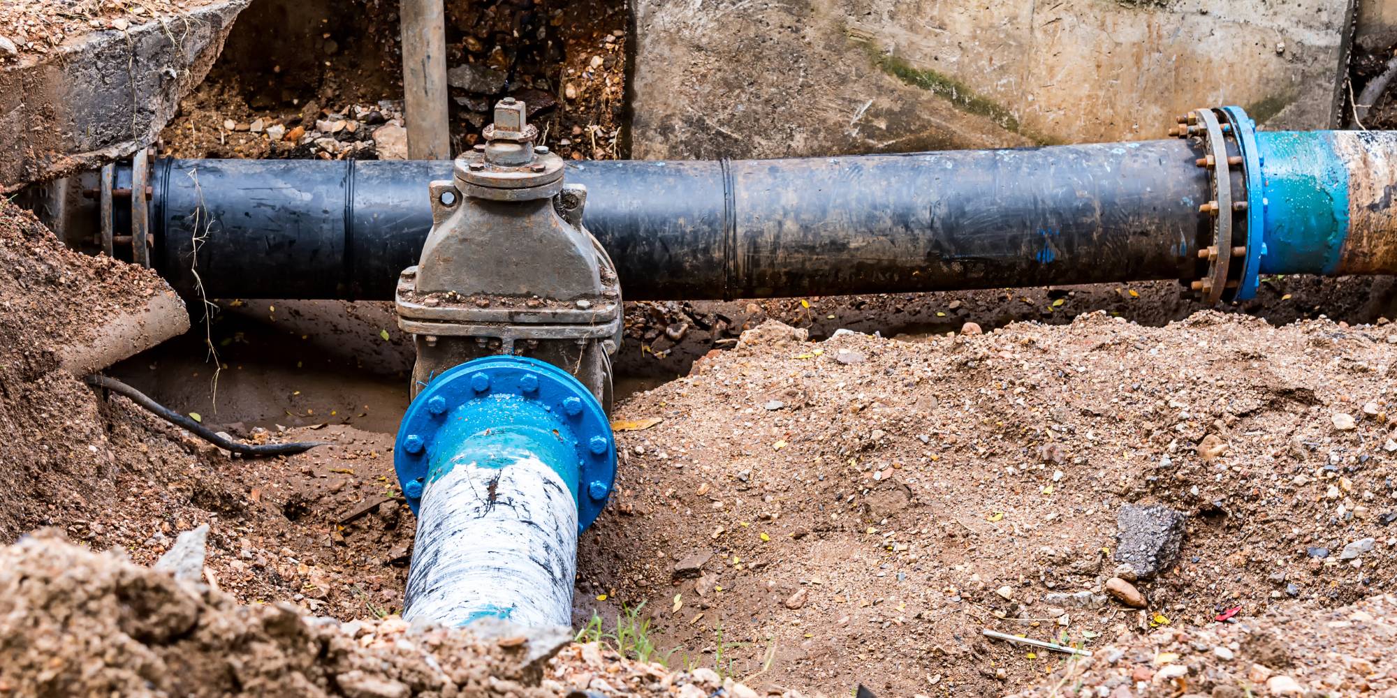 Large black and blue water pipes with a valve are partially buried in dirt at a construction or repair site, with surrounding soil and concrete walls visible.