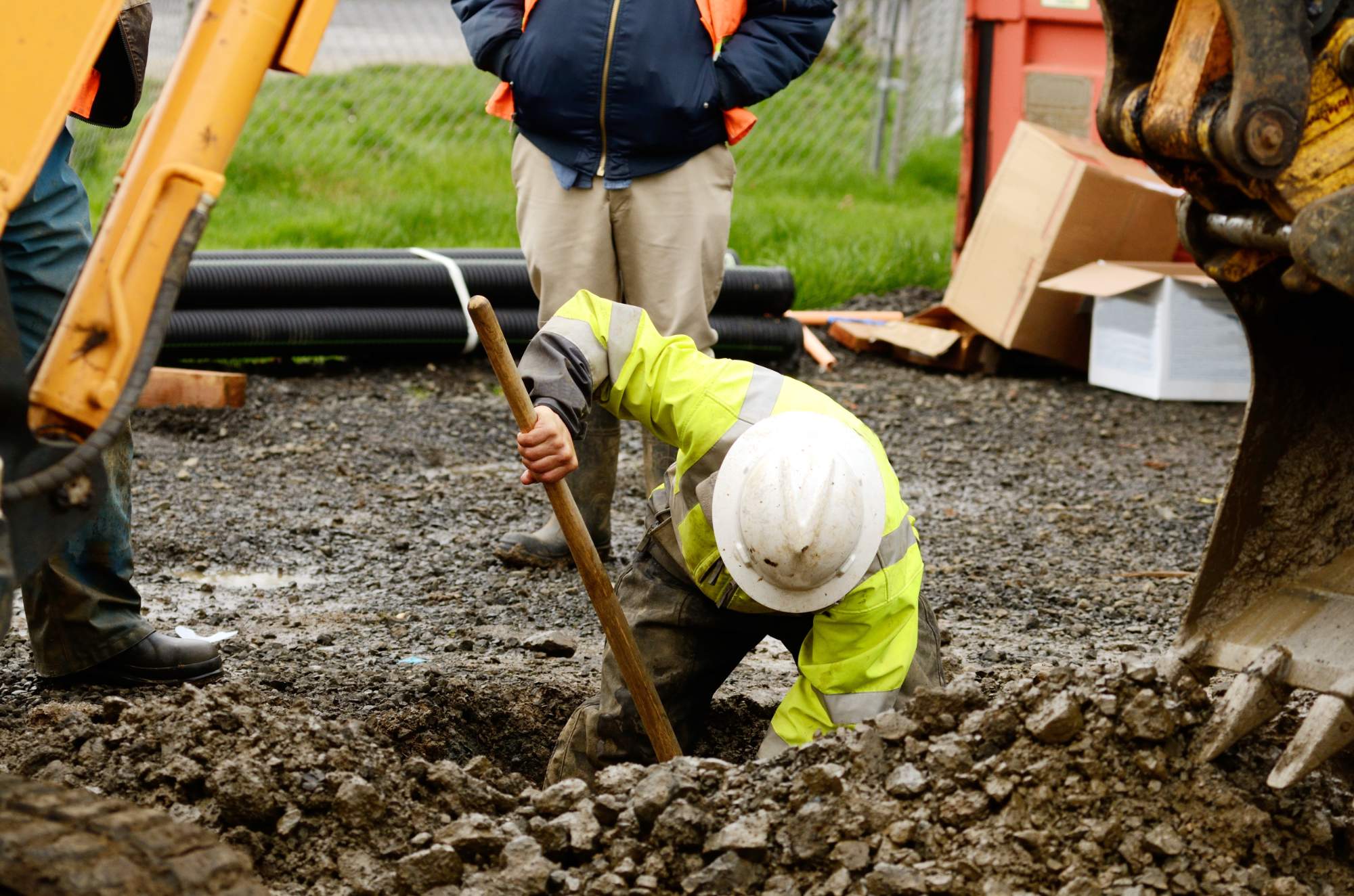 A construction worker in a yellow safety jacket and white hard hat digs into muddy ground with a shovel, while two other people stand nearby, one wearing a navy jacket and orange vest. Construction equipment is visible.