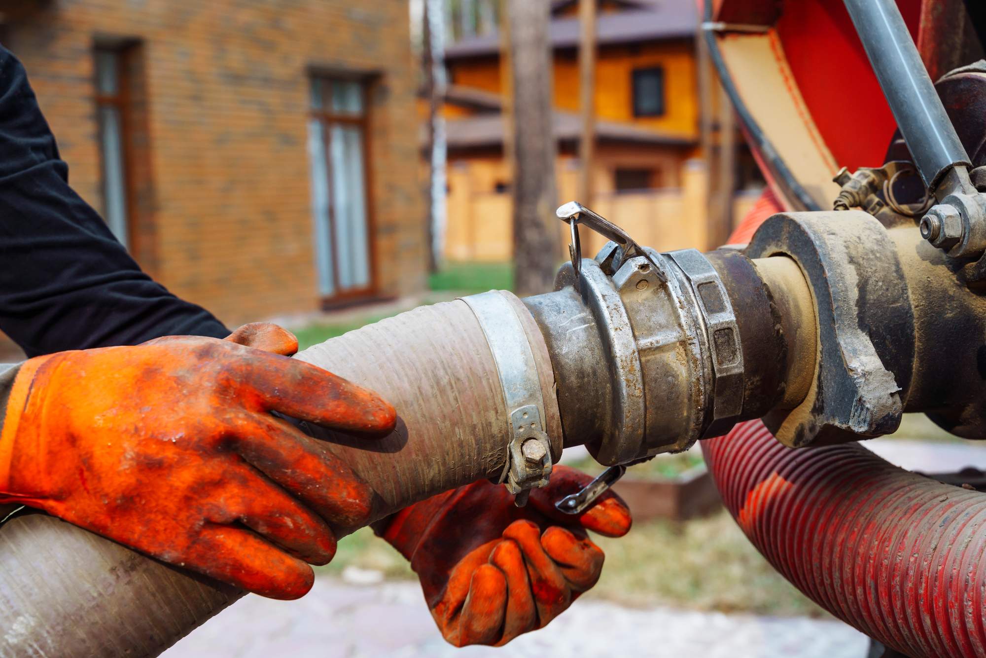 A person wearing orange gloves connects a large hose to a sewage or septic truck, with houses and trees visible in the background.