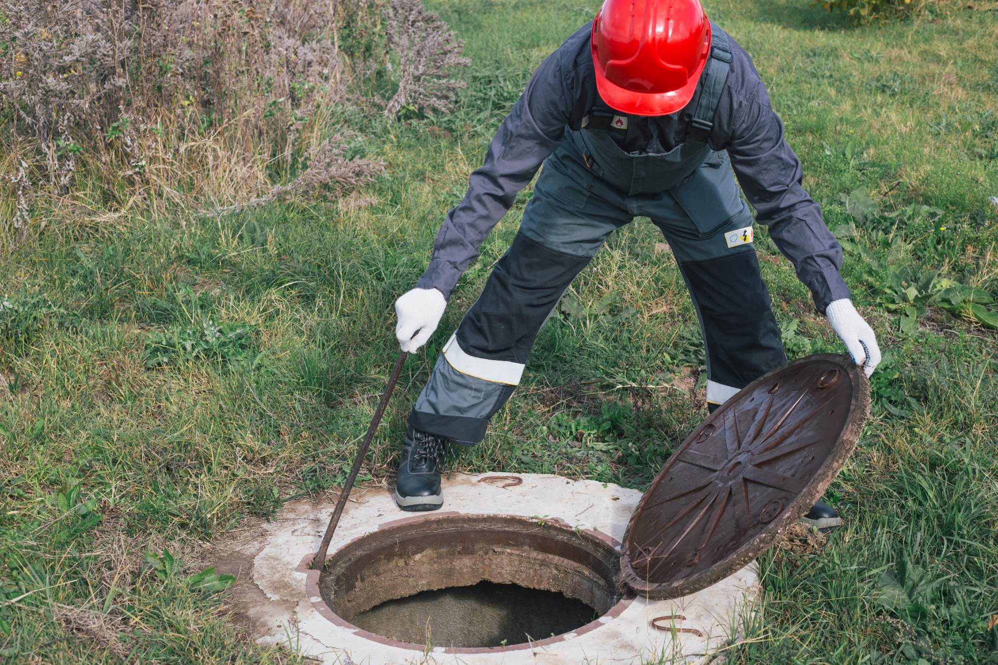 A worker in protective clothing and a red helmet lifts a manhole cover with a tool in a grassy outdoor area.
