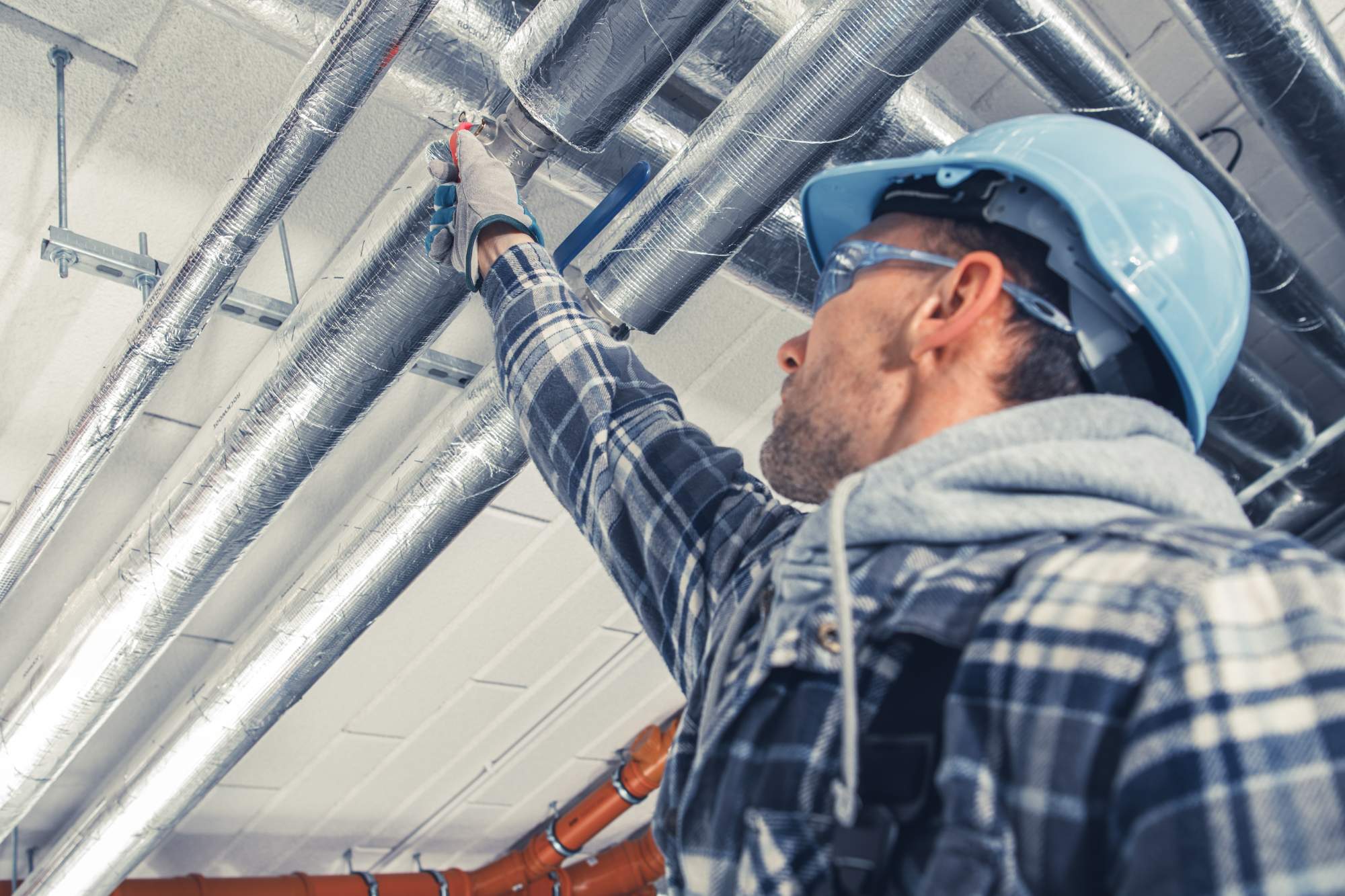 A construction worker in a plaid shirt, safety helmet, and goggles inspects or repairs overhead metal pipes on a ceiling, reaching up with one gloved hand.
