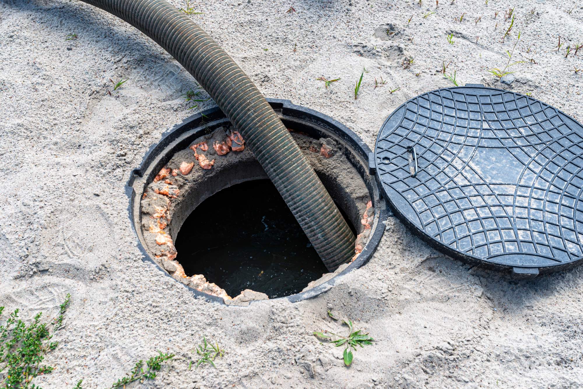 A large hose is inserted into an open manhole surrounded by sand, with the round manhole cover lying to the side on the ground.