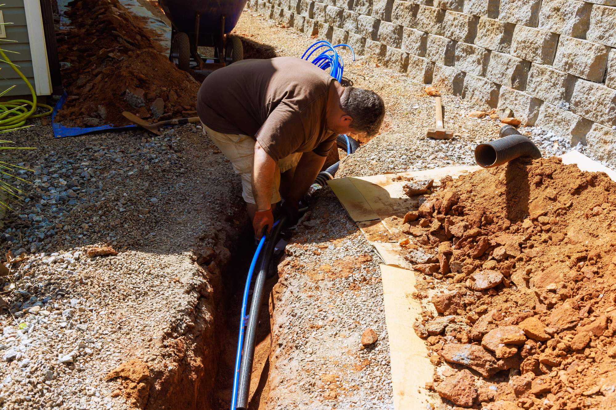 A man bends over a narrow trench, installing blue pipes in the ground next to a stone retaining wall, with soil and tools scattered nearby on a sunny day.