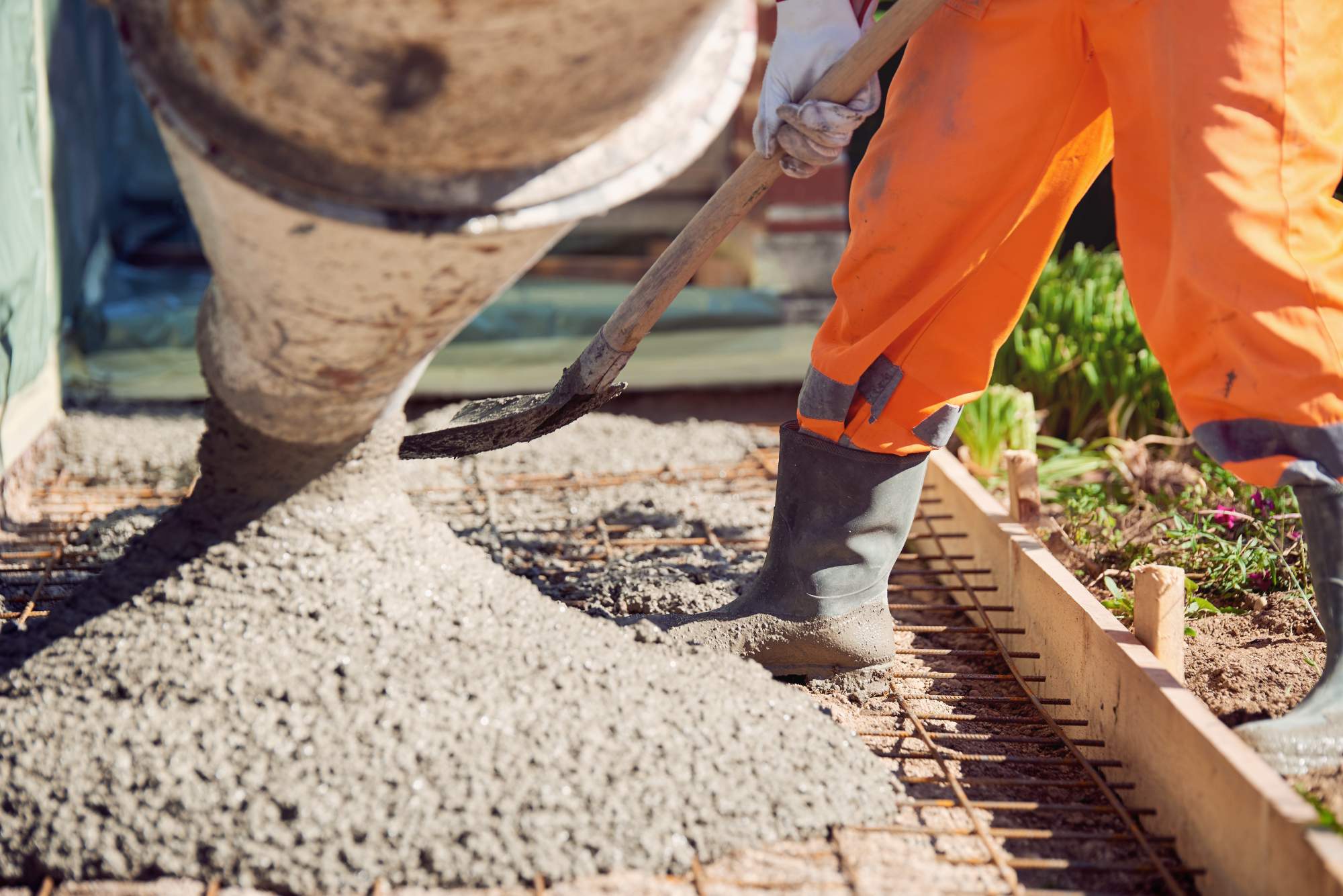 A construction worker wearing orange pants and rubber boots spreads freshly poured concrete with a shovel onto a rebar-reinforced surface at a building site.