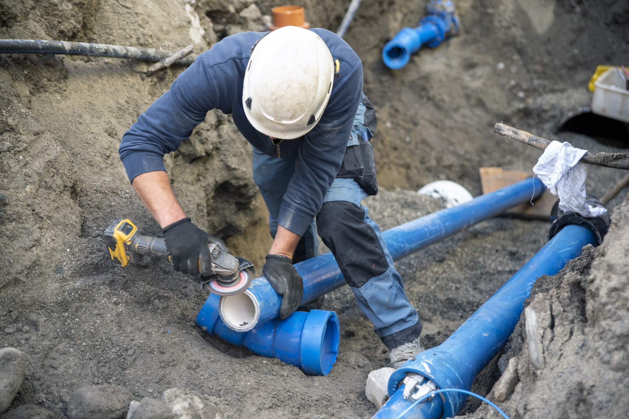A construction worker wearing a helmet and gloves uses a power tool to cut a blue plastic pipe in a trench surrounded by soil and other pipes.