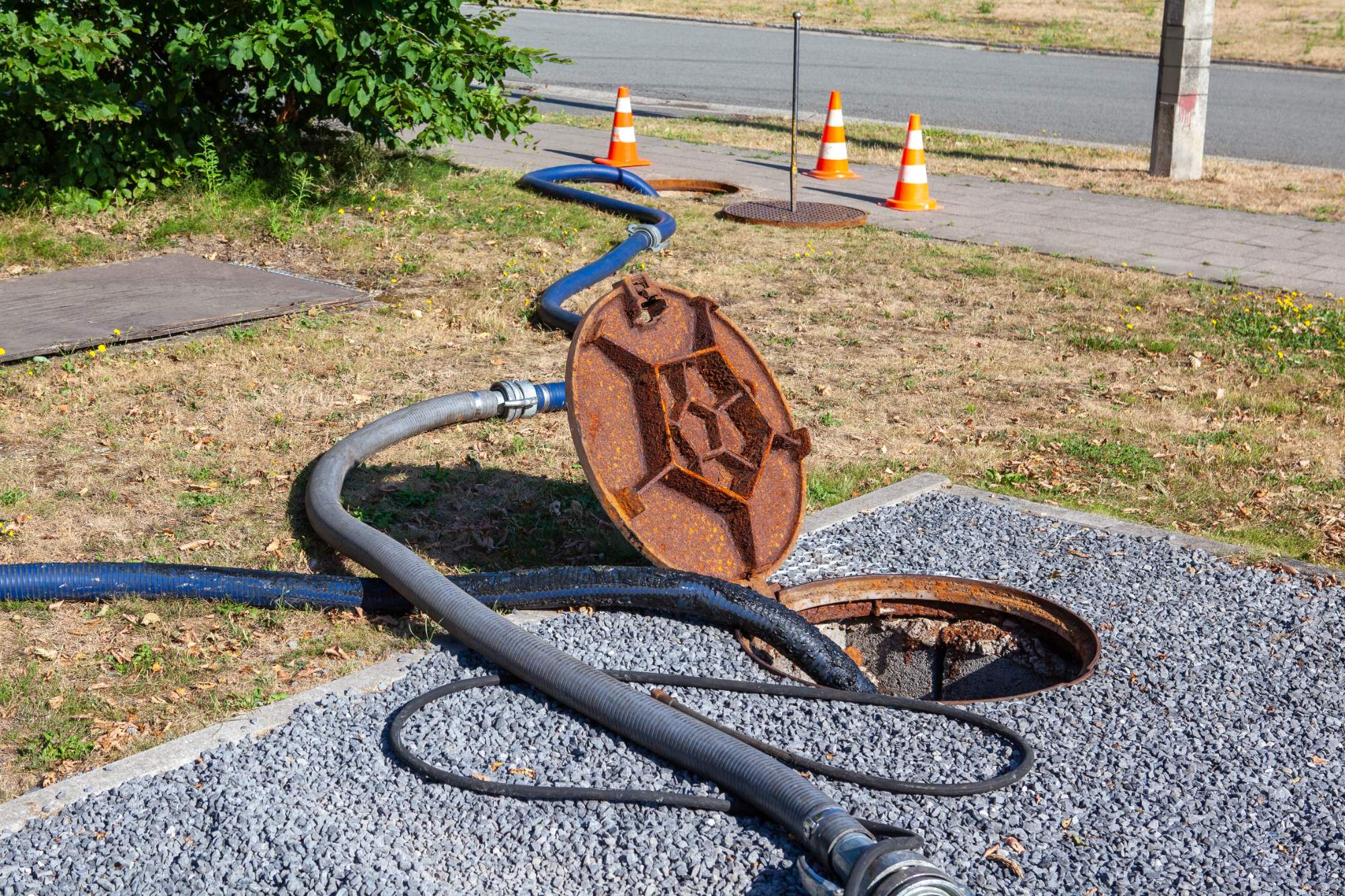 A rusty manhole cover is open with several hoses leading into the sewer. Orange traffic cones mark the area on a grassy roadside, indicating ongoing maintenance work. Gravel and pavement are visible around the manhole.