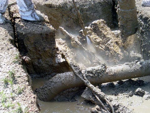 Workers in protective gear handle a large, muddy pipeline partially buried in wet soil, with water spraying around as they clean or excavate the area.