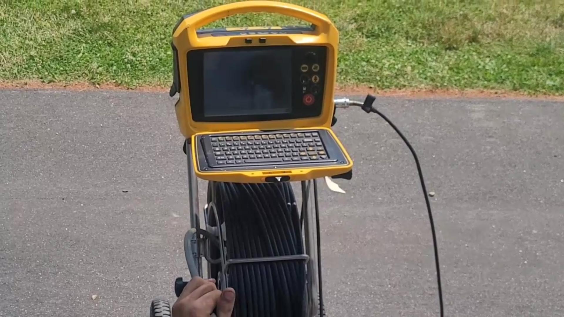A yellow portable inspection camera system with a screen and keyboard is mounted on a wheeled frame, with a coiled black cable attached. It is set up on a paved outdoor surface near a grassy area.