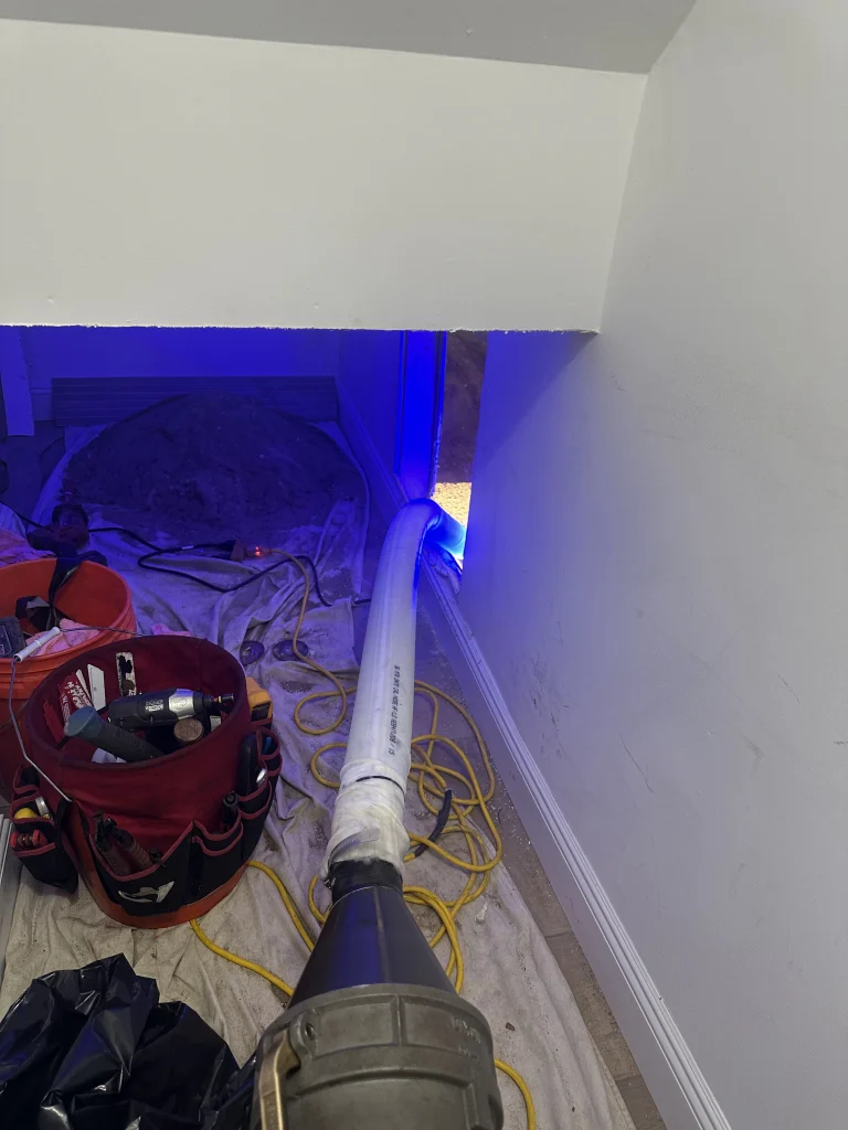 A large hose extends from a machine into a small, unfinished room with a dirt floor and blue lighting. Tools, a red tool bag, and extension cords are scattered on a tarp covering the floor.